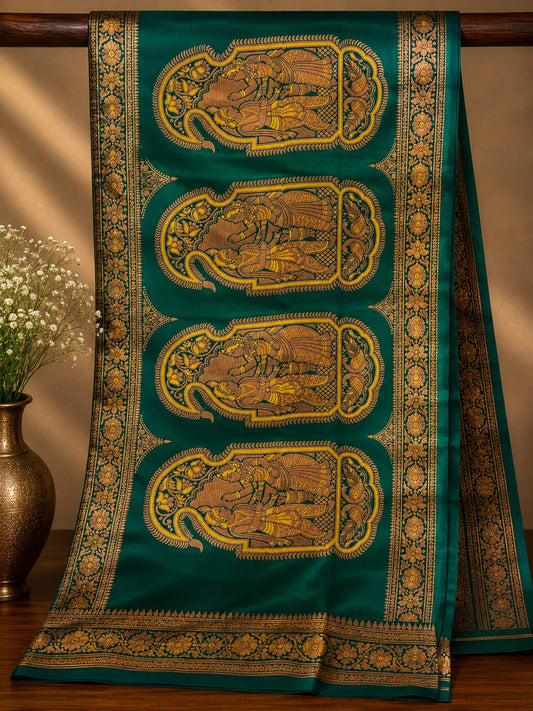 A dark teal green silk saree with four large gold and brown elephant motifs and ornate borders, draped elegantly on a wooden stand next to a vase of white flowers.