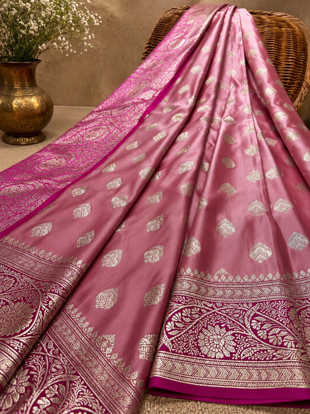 A pink Banarasi silk saree with intricate silver zari brocade work, draped elegantly over a wicker chair next to a brass vase with white flowers.