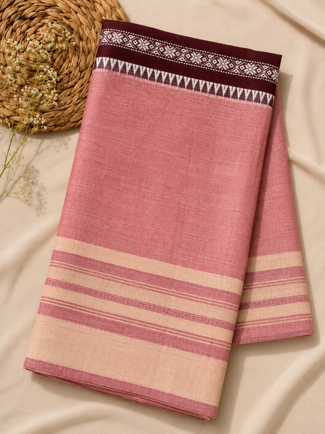 A folded pink sari displayed on a cream-colored fabric background with a woven straw hat and dried flowers as props. The fabric features a dark maroon border with white geometric patterns and horizontal stripes in pink and cream.